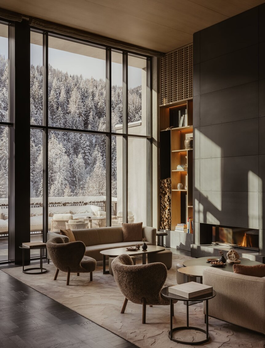 The double-height lounge at Aman Rosa Alpina, with a fireplace and snow-covered forest beyond the windows