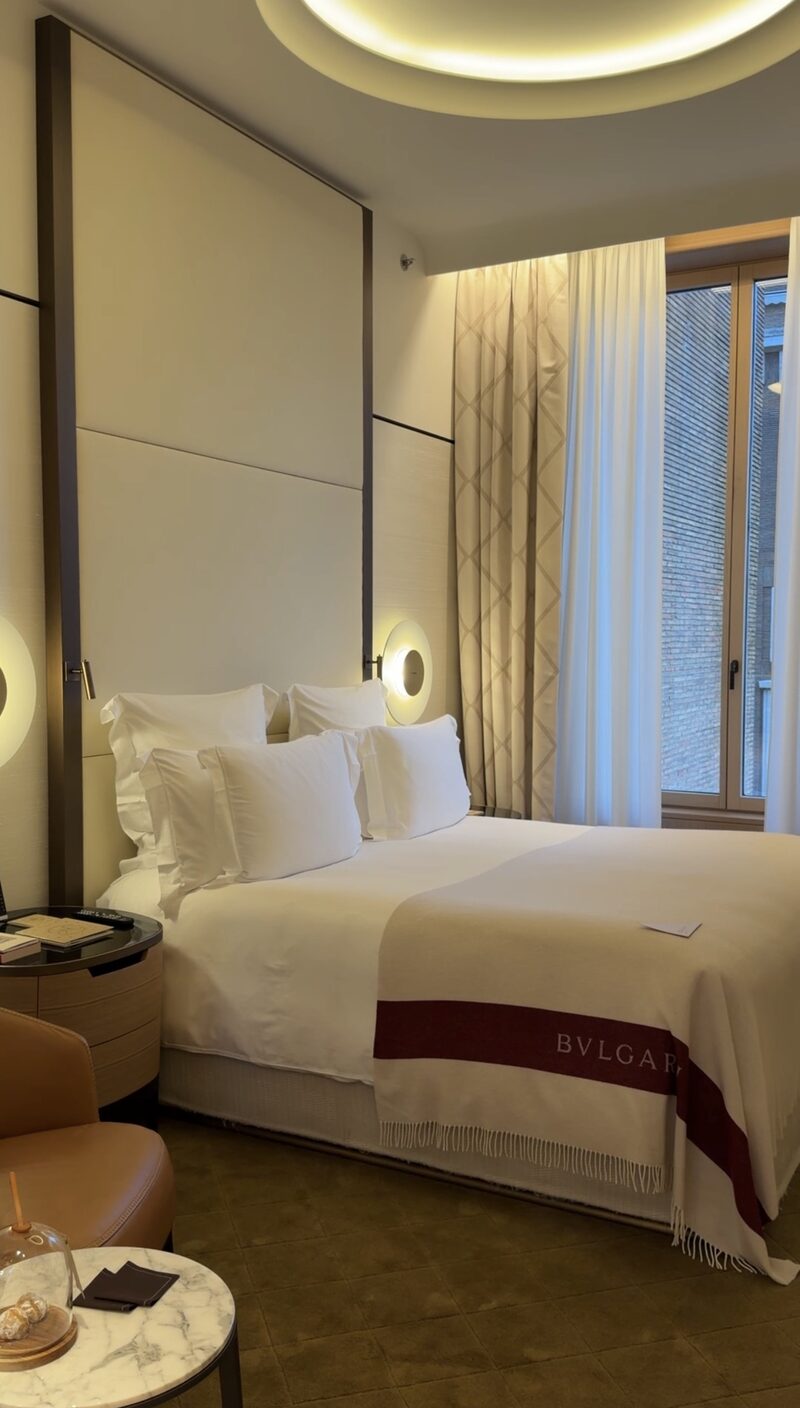 A Bulgari Hotel Roma bedroom with a tall upholstered headboard, round brass reading lights and a Bvlgari throw on the bed
