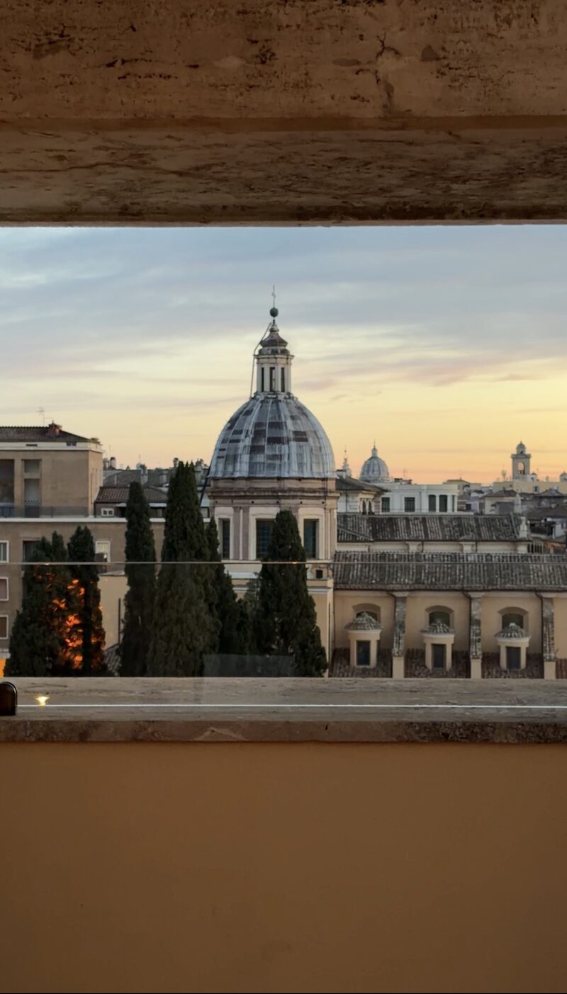 A church dome and cypress trees framed by a travertine window at the Bulgari Hotel Roma at dusk