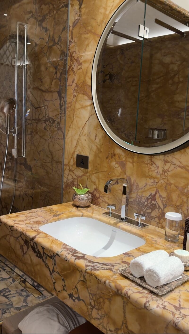 A Bulgari Hotel Roma bathroom vanity in golden veined marble with a round mirror and a glass shower
