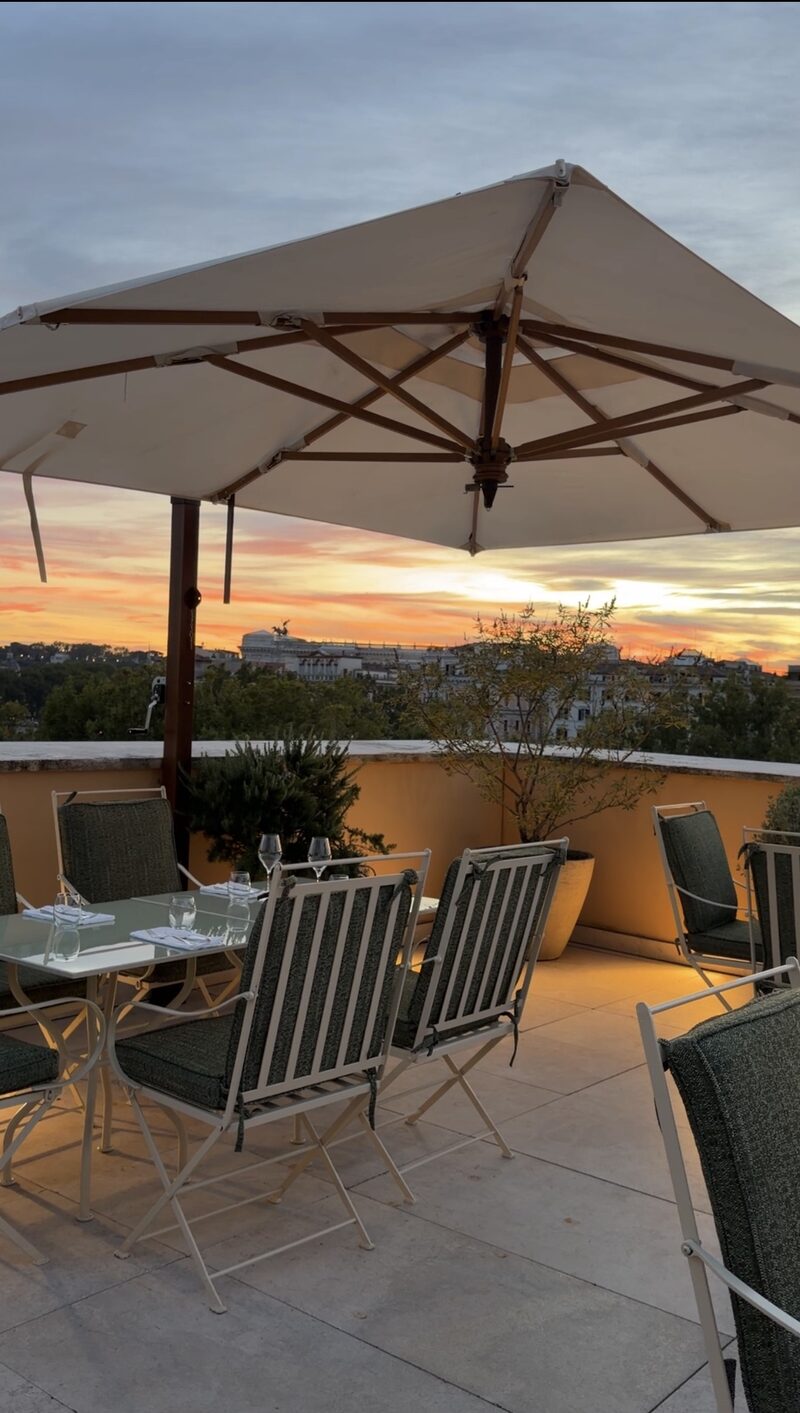 A Bulgari Hotel Roma terrace table under a white umbrella at sunset, looking over trees and rooftops