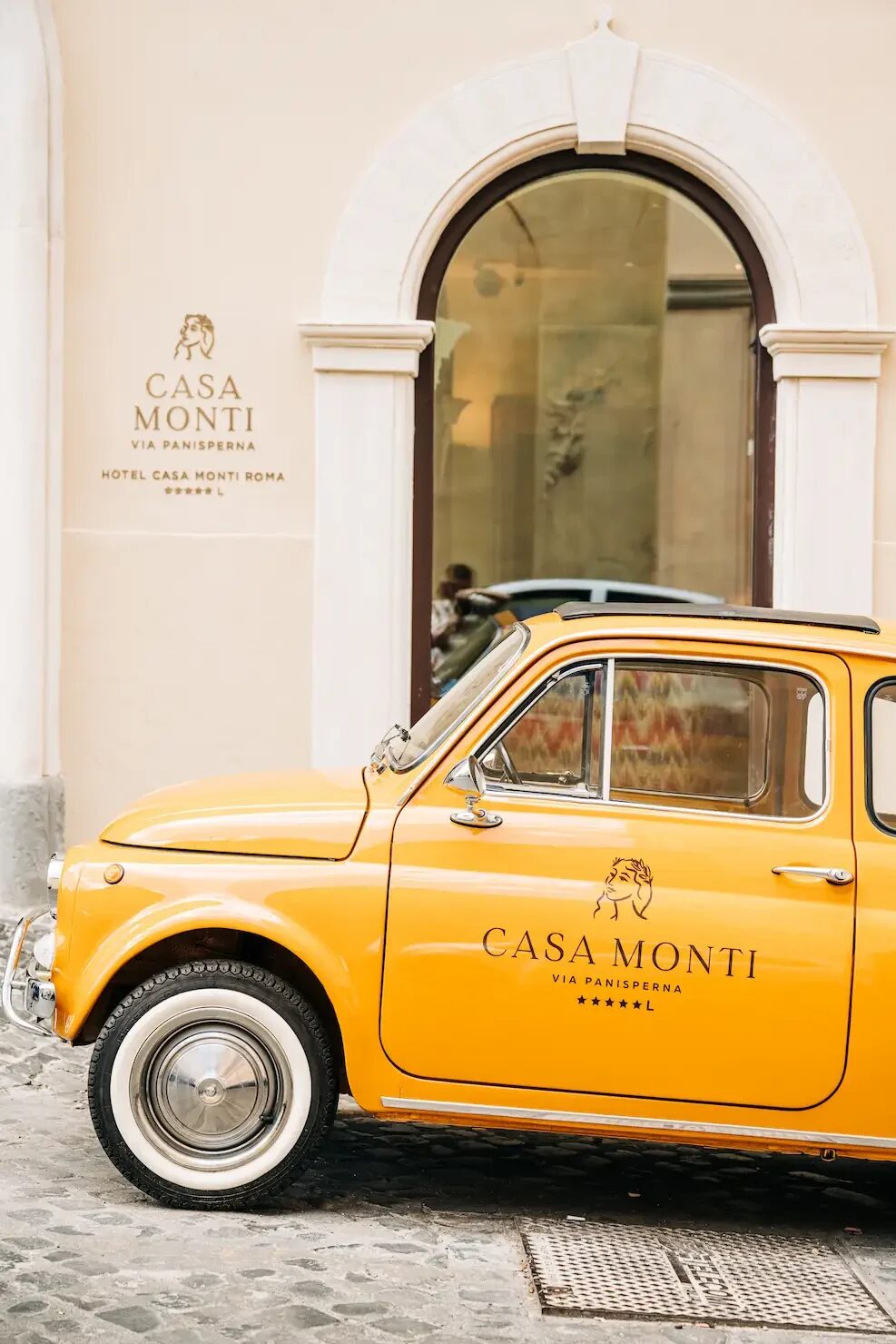 A yellow vintage Fiat 500 with the Casa Monti logo parked outside the hotel&rsquo;s arched entrance on Via Panisperna
