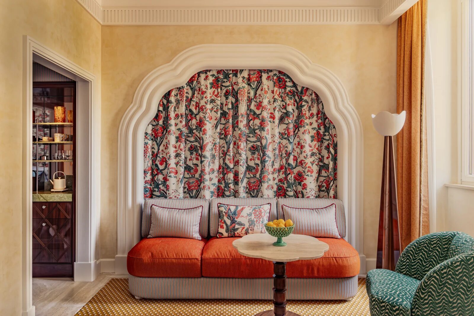 A Casa Monti room with an orange banquette under a scalloped arch of bird-print fabric, a green zigzag armchair and a marble-topped table
