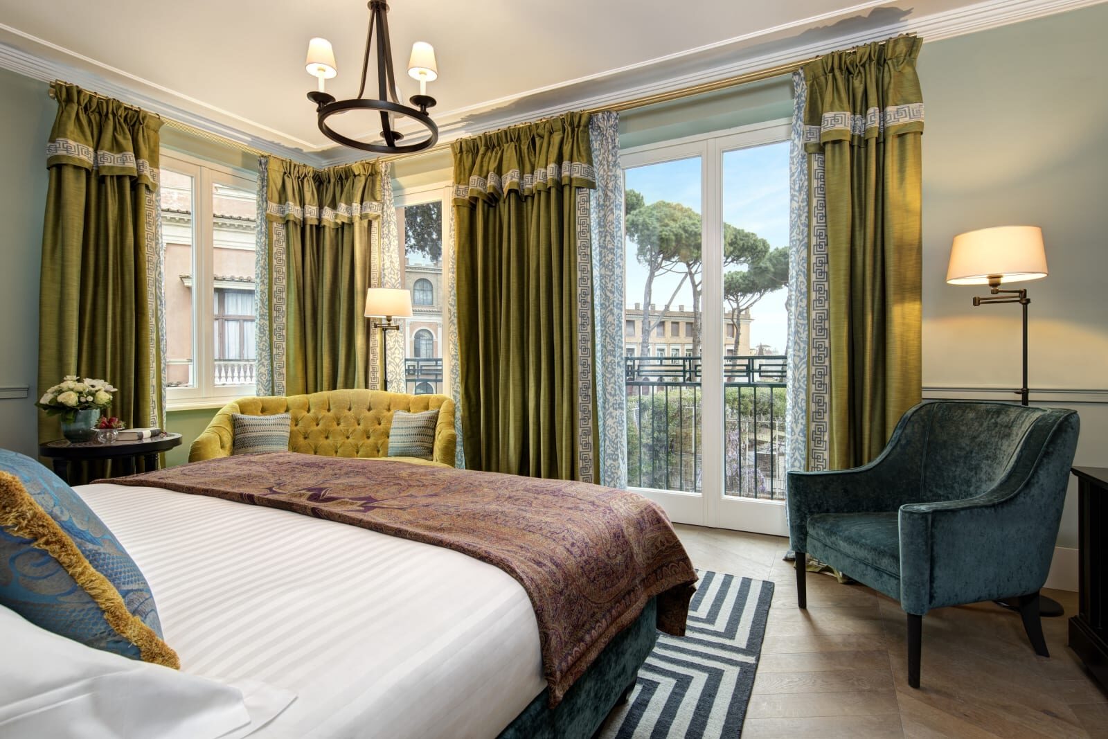 A Hotel de la Ville bedroom with green velvet curtains, a yellow loveseat and a balcony looking out to umbrella pines