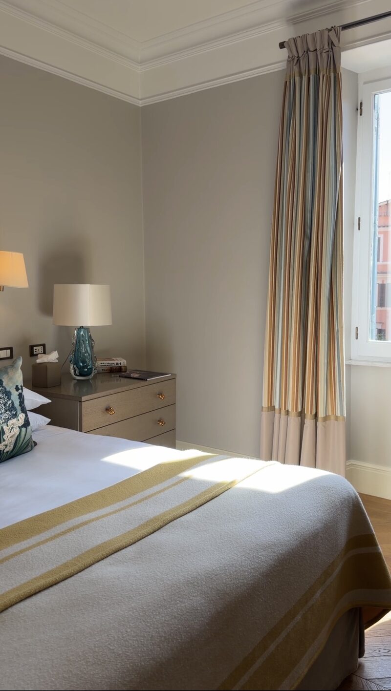 A sunlit Hotel de Russie bedroom with soft gray walls, striped curtains and a window onto the rooftops
