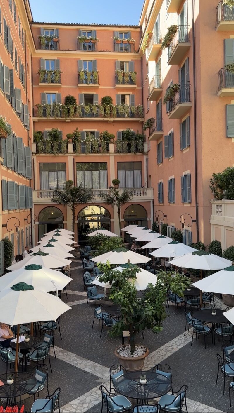 Hotel de Russie’s large courtyard, with white umbrellas over tables and salmon-pink walls with blue-gray shutters and flowering balconies