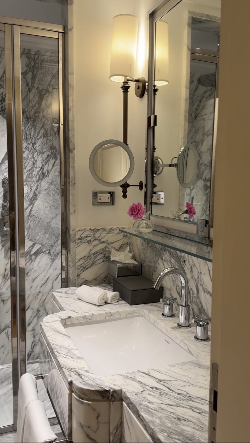 A Hotel de Russie bathroom in white and gray veined marble, with a sconce, magnifying mirror and glass shower