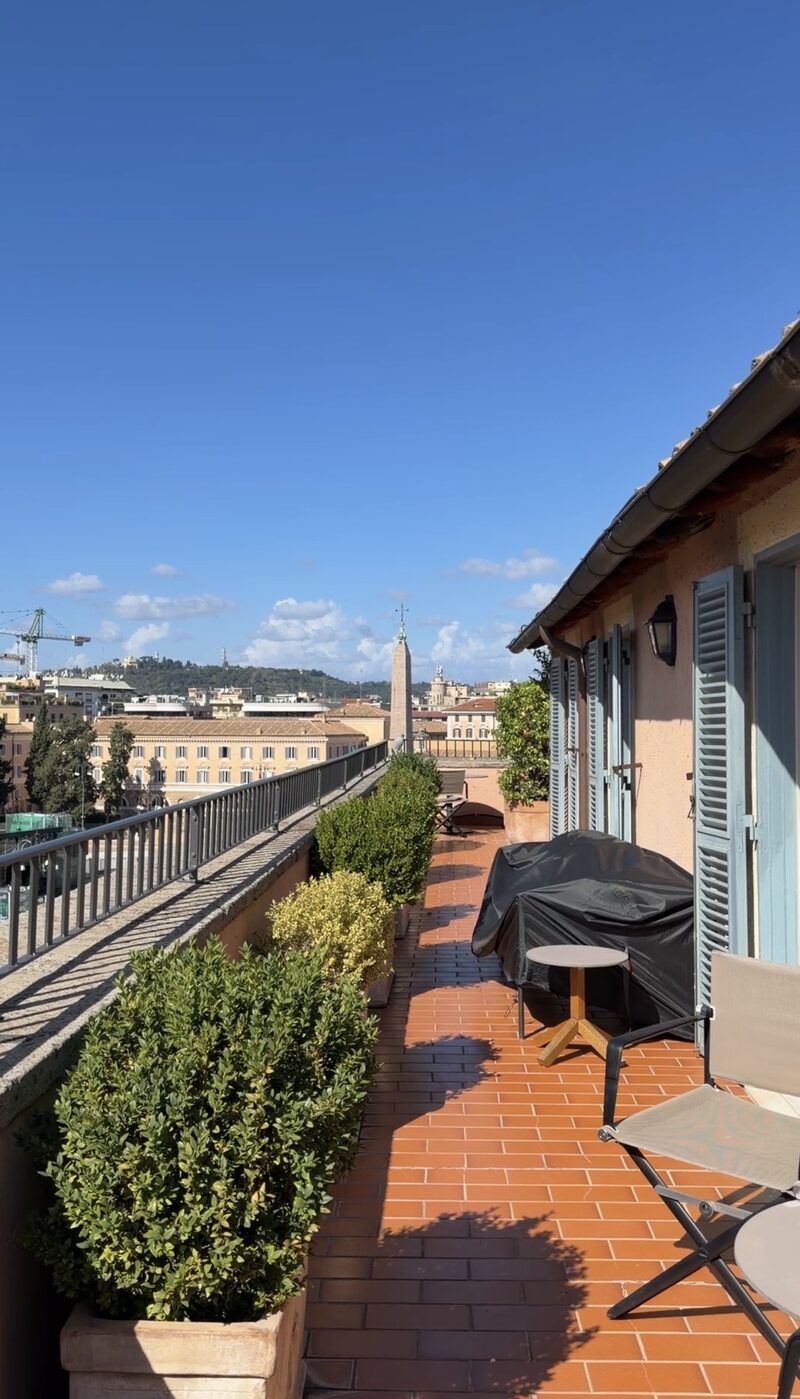 A Hotel de Russie suite terrace with boxwood planters and blue shutters, looking over Piazza del Popolo’s obelisk to the hills