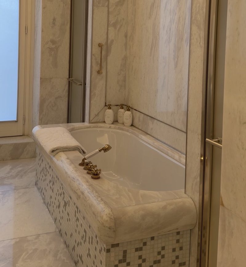 A white marble Hotel Eden bathroom with a deep soaking tub and brass fixtures