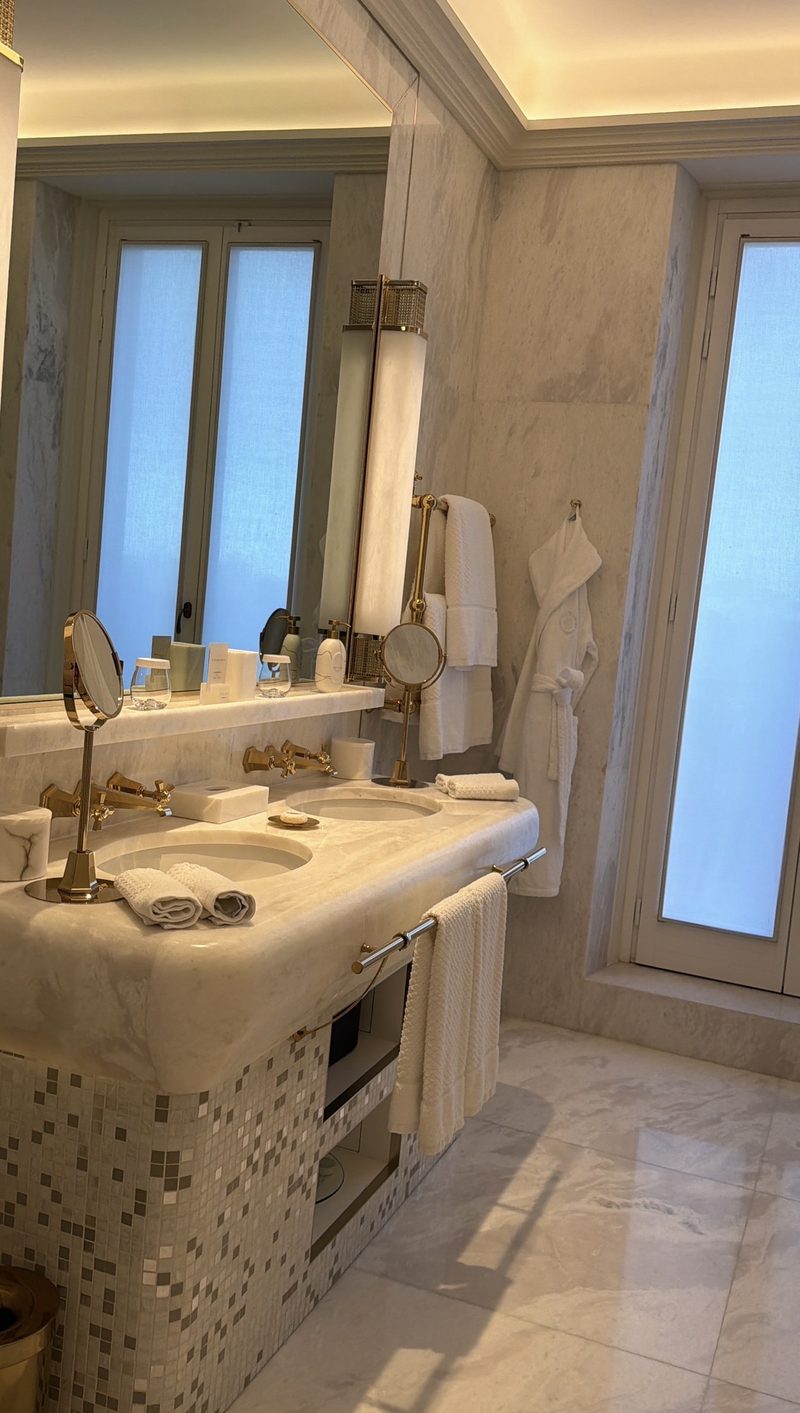 A Hotel Eden bathroom with a double marble vanity, brass fixtures and a robe on the wall