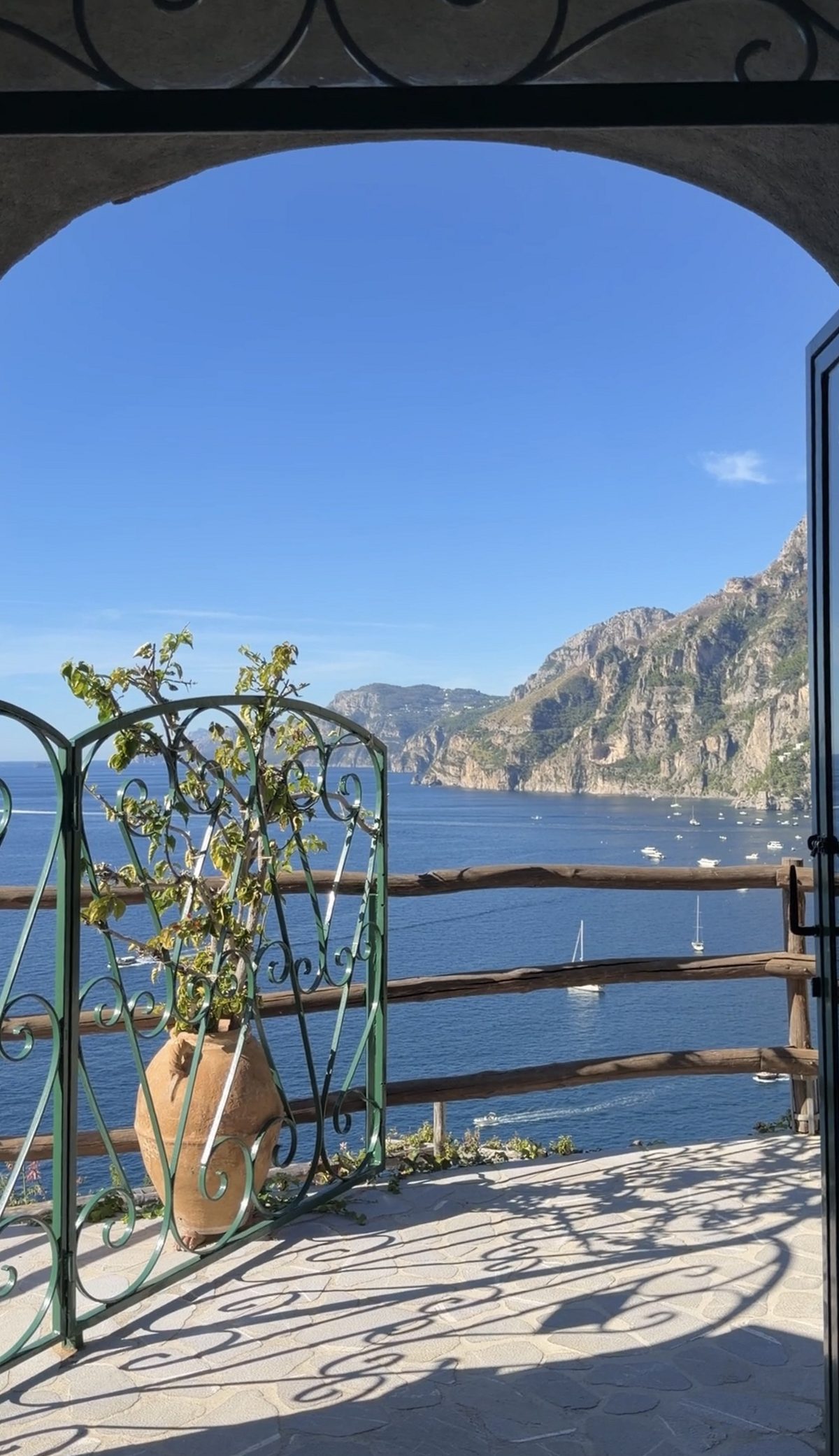 A green iron gate under a stone arch opening onto a terrace above the bay, with boats and the cliffs of the Amalfi Coast