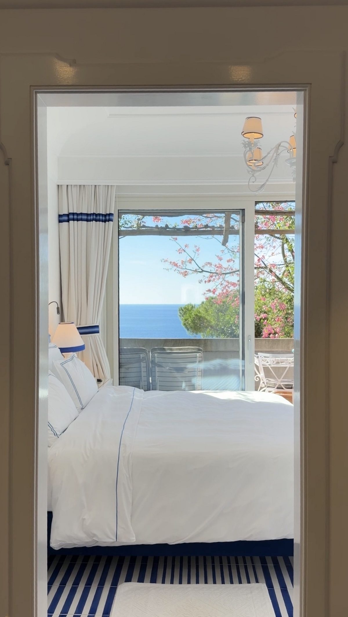 Through a doorway to a white bed with blue piping and a terrace door framing the sea and pink blossoms