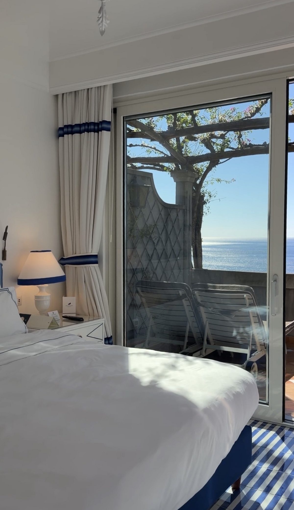 A bedroom with blue-striped tiled floors and sliding doors to a private terrace with loungers above the sea