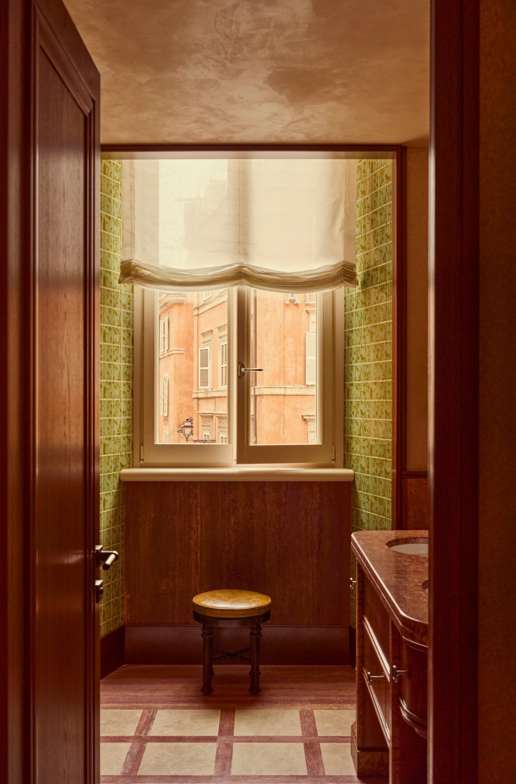 A bathroom with green hand-painted tiles and a window onto ochre Roman facades