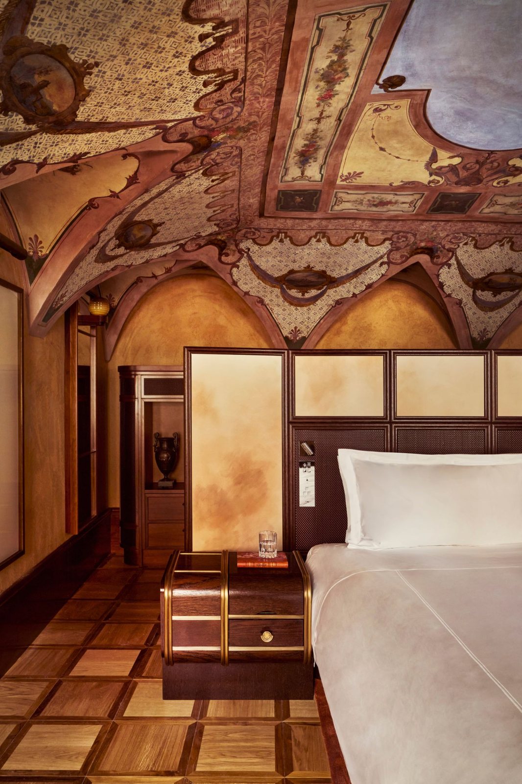A bedroom beneath a painted vaulted ceiling, with a brass-banded nightstand and parquet floor