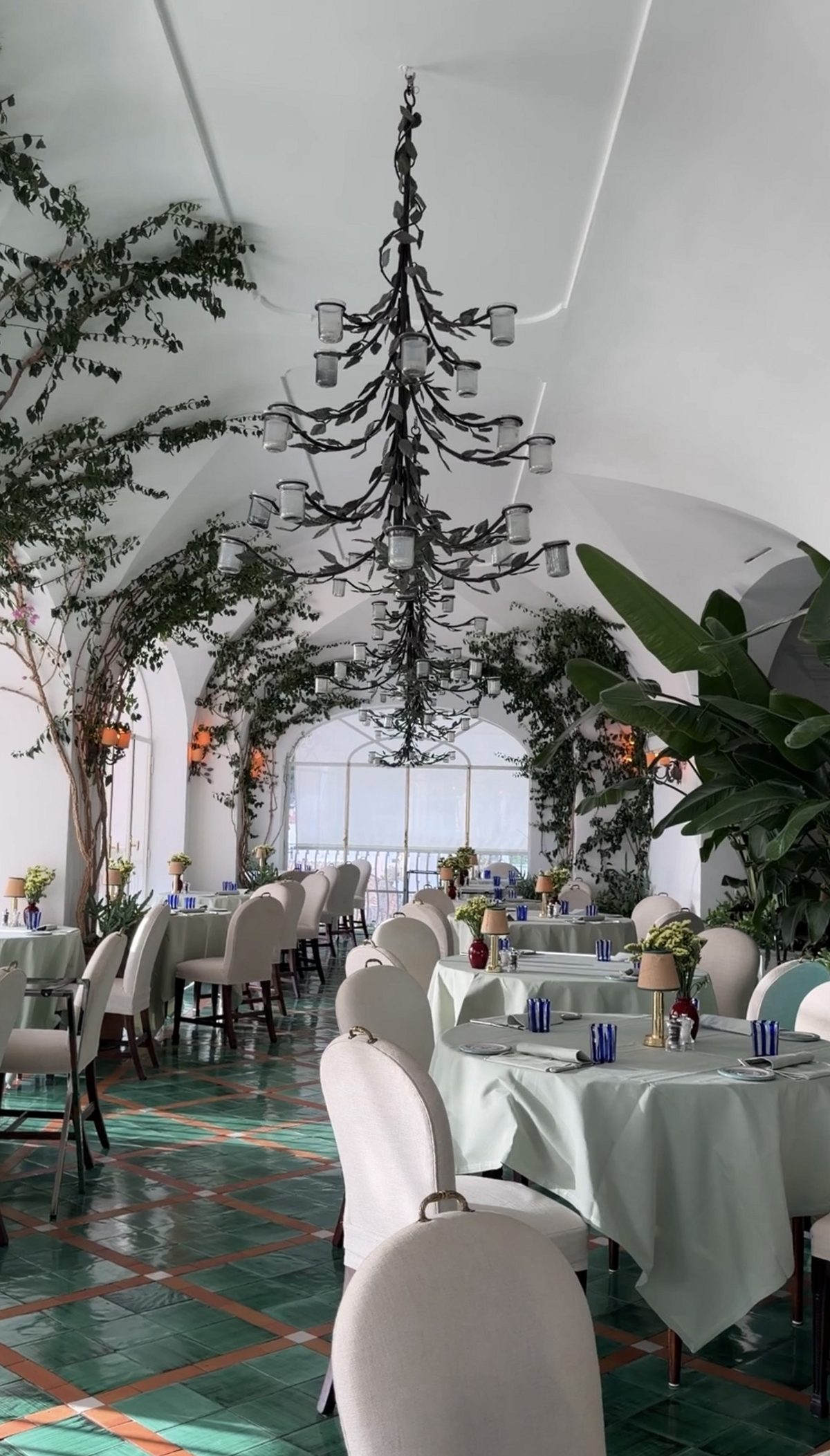 A vaulted white dining room at Le Sirenuse with iron chandeliers, climbing vines and green tiled floors