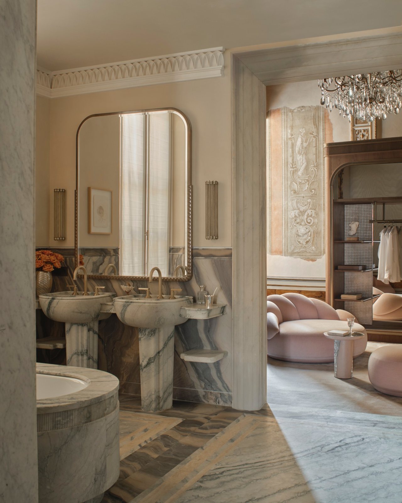 Twin marble pedestal basins in a suite bathroom, opening onto a dressing room under a crystal chandelier