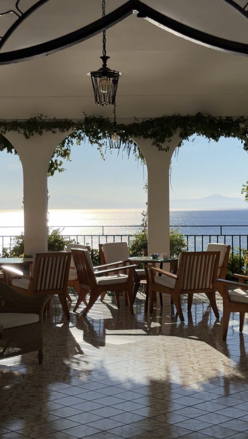 Wooden armchairs under white arches draped in vines, looking out to the glittering sea at Hotel Santa Caterina