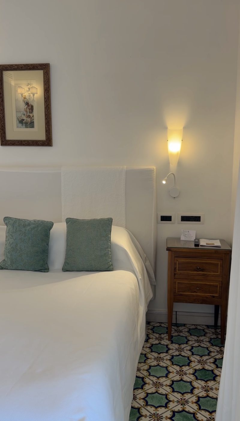 A white bed with sage-green cushions, a walnut nightstand and majolica tile floor at Hotel Santa Caterina