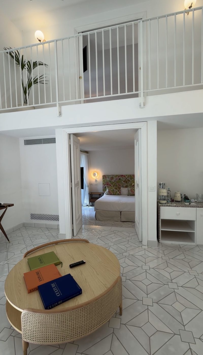 A double-height Casa Marchesa living room with a gallery above, a rattan coffee table and a bedroom through double doors