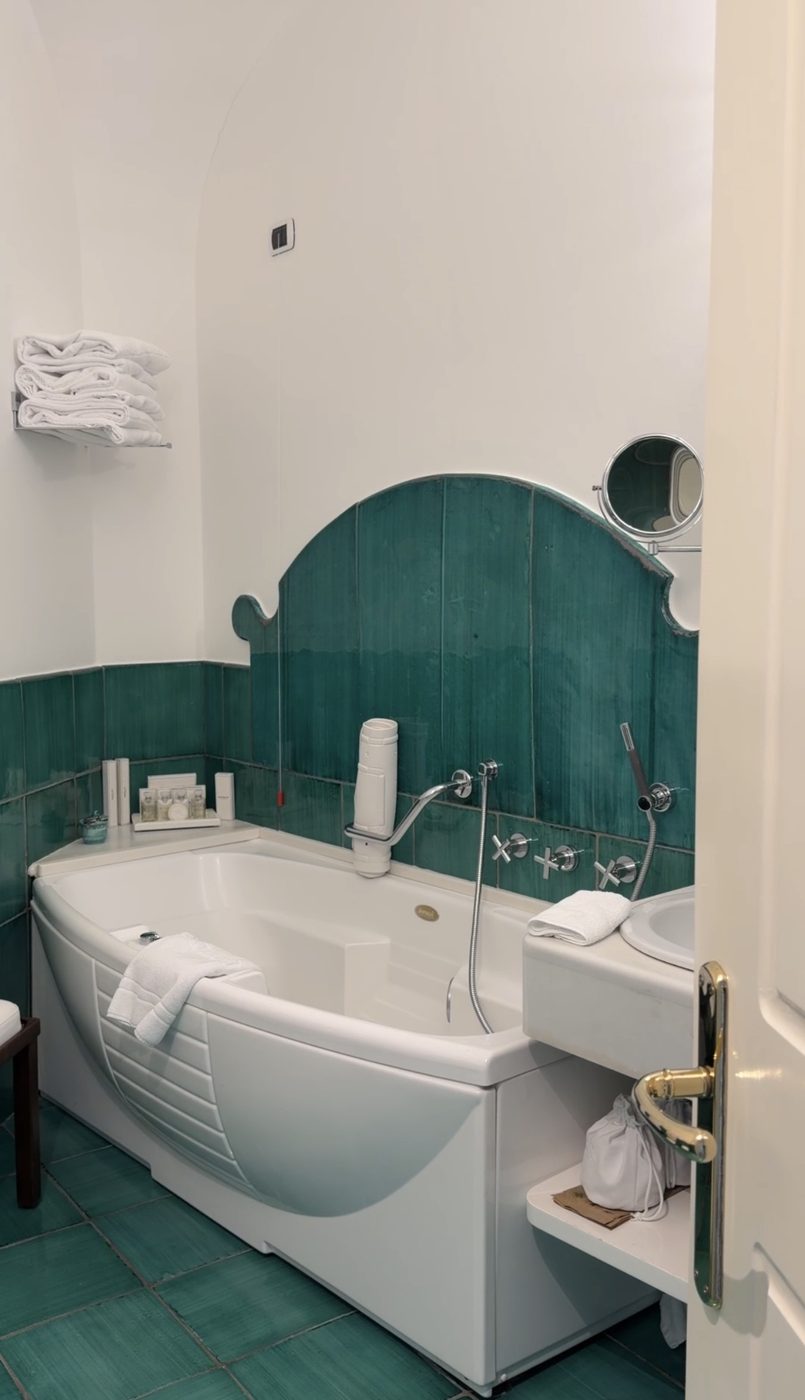 A bathtub set against hand-glazed green tiles with a scalloped tiled headboard at Hotel Santa Caterina