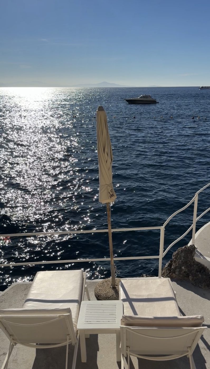Two white loungers and a furled umbrella at the railing over a sparkling sea, a boat moored offshore