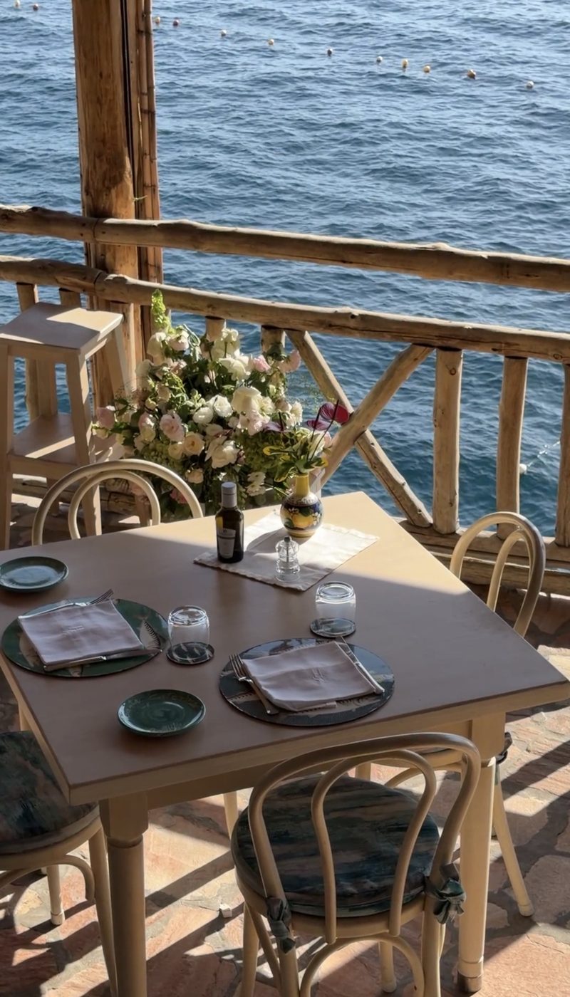 A table set with olive oil, a lemon-painted vase and a bouquet of roses beside a rustic railing above the sea