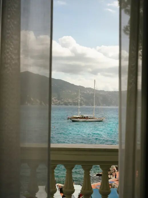 View through sheer curtains from a balustraded balcony at Grand Hotel Miramare to a sailboat anchored in the bay