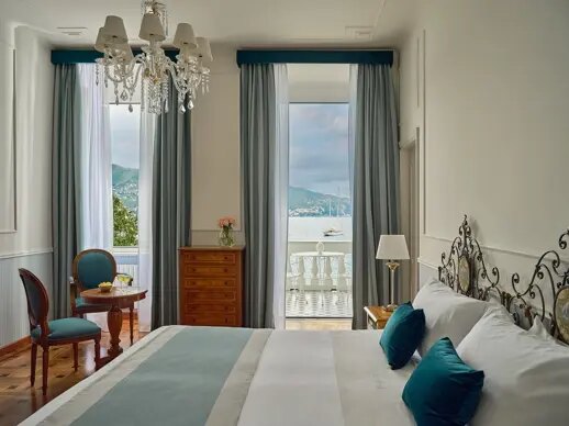 A restored sea-view room at Grand Hotel Miramare with a chandelier, an iron bedframe and French doors open to a balcony over the bay