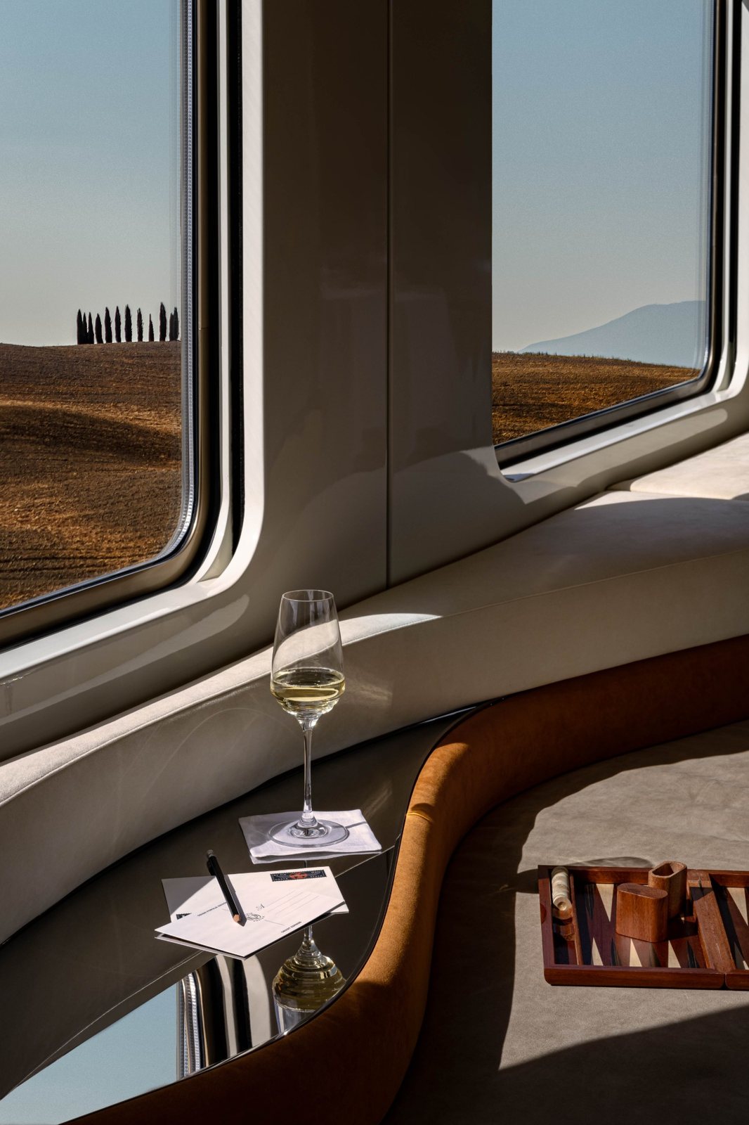 A glass of wine, postcards and a backgammon board on a curved sofa beside the windows of the bar car